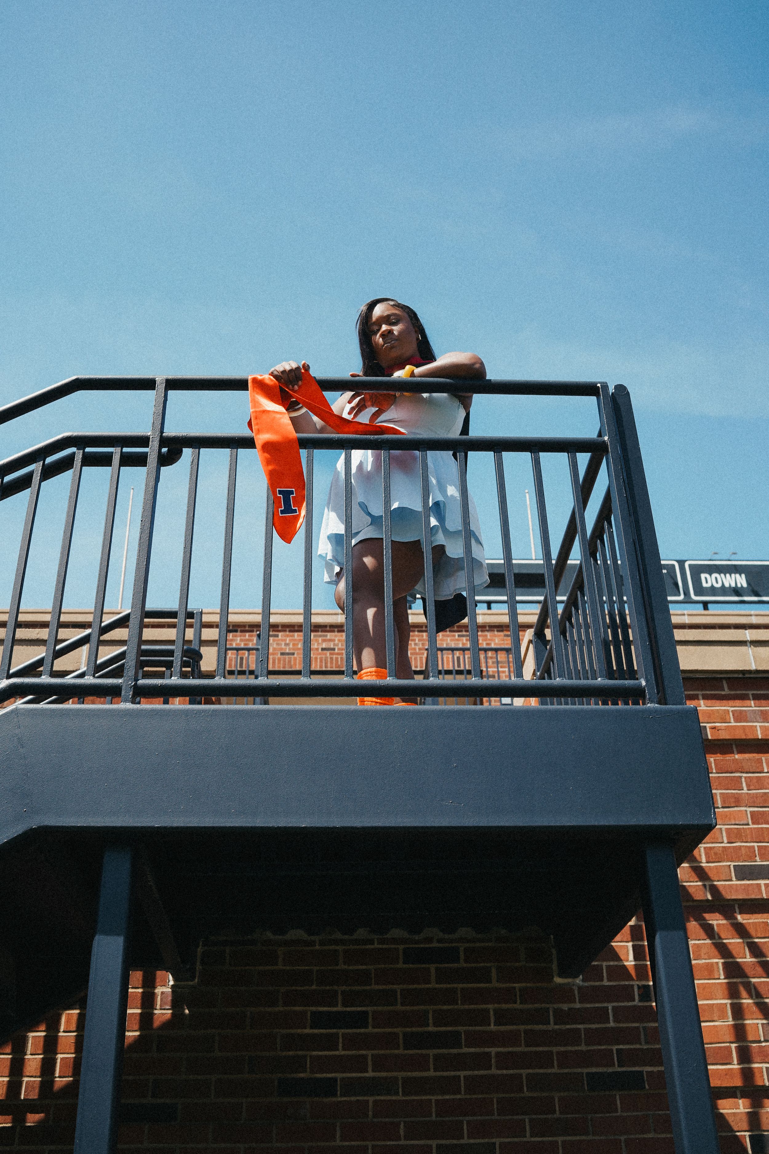 Orange dress contrasts against dark metal balcony railing under blue sky. graduation