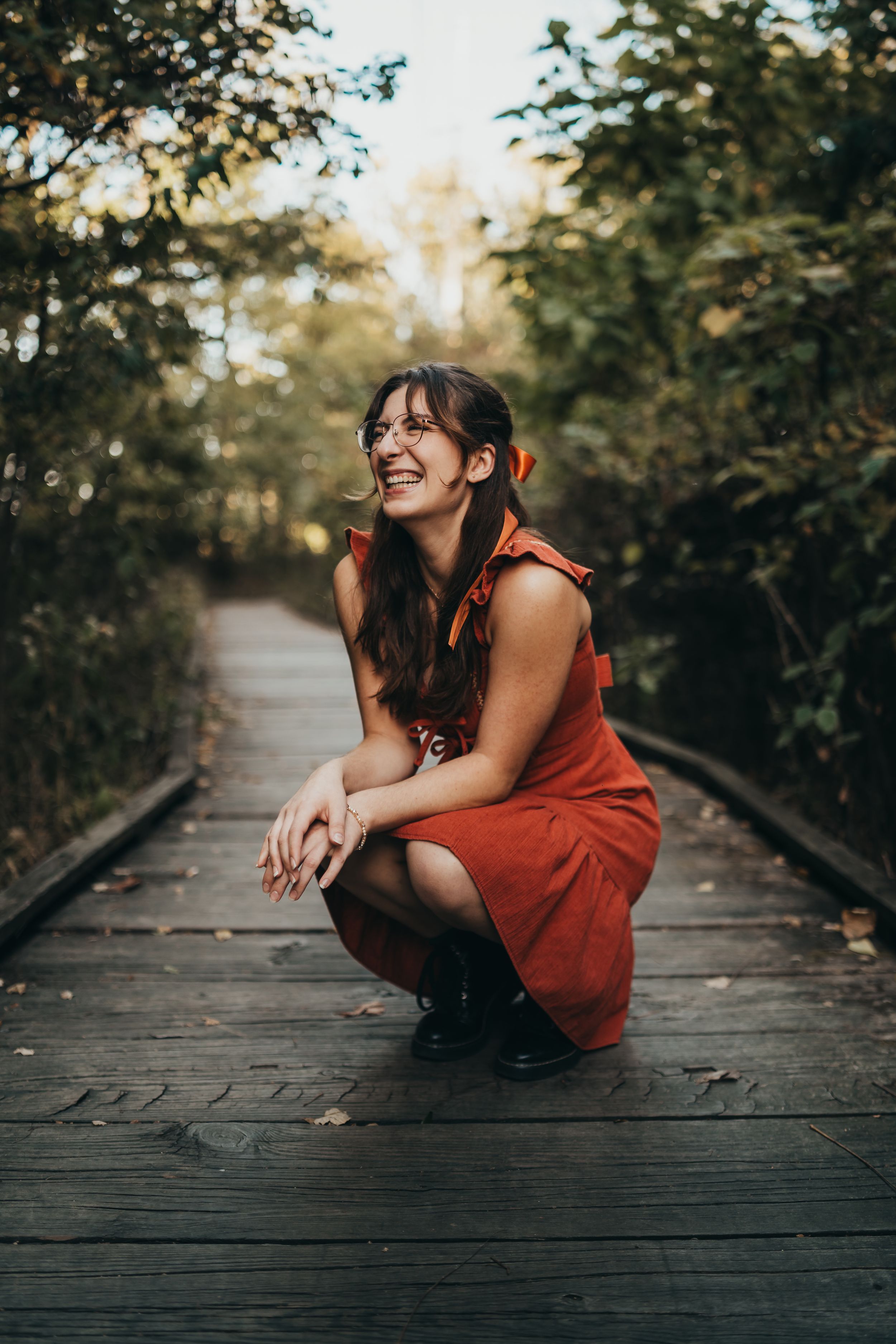 Woman in red dress crouching on wooden boardwalk surrounded by trees.