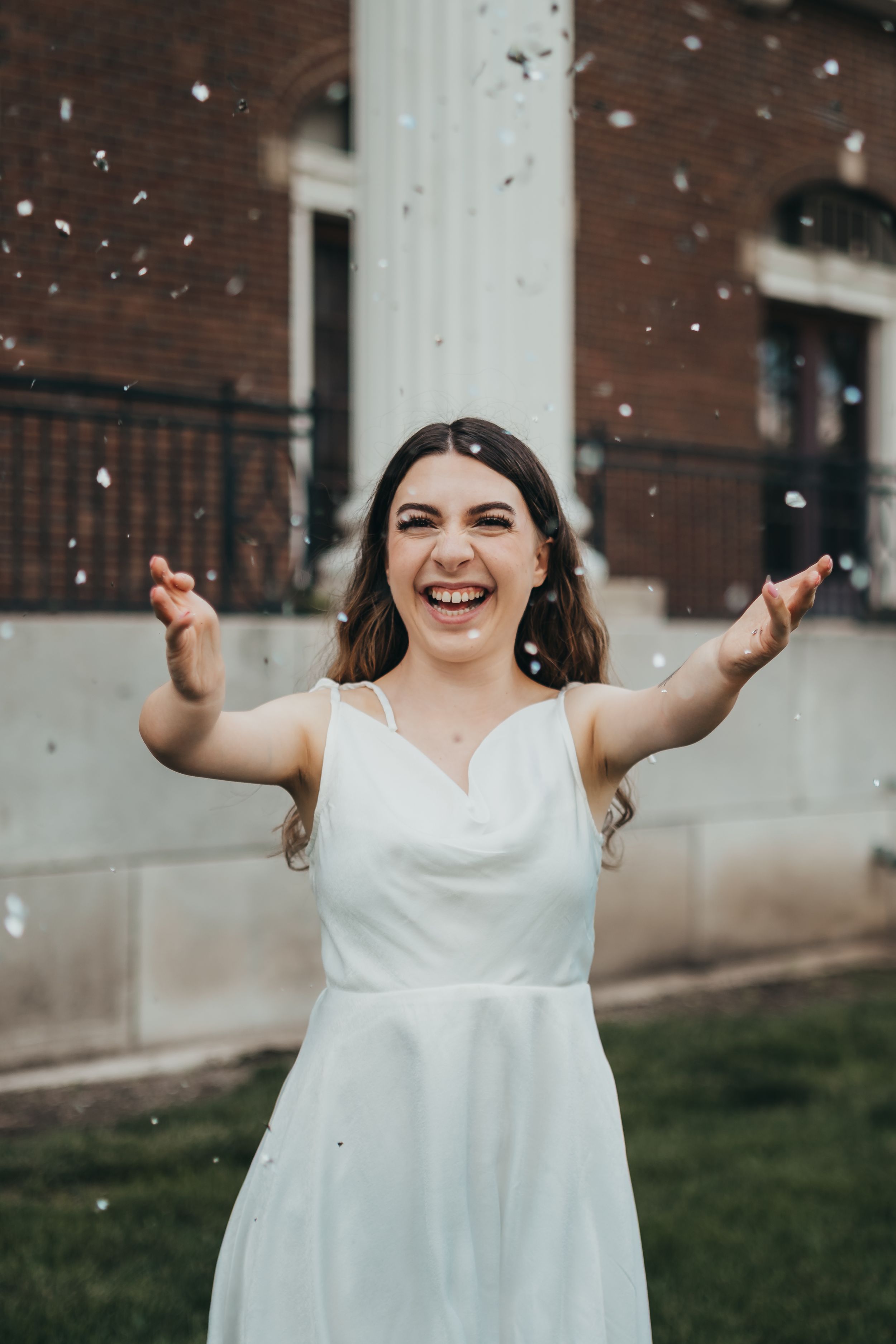 Person in white dress celebrating with confetti against brick wall background.