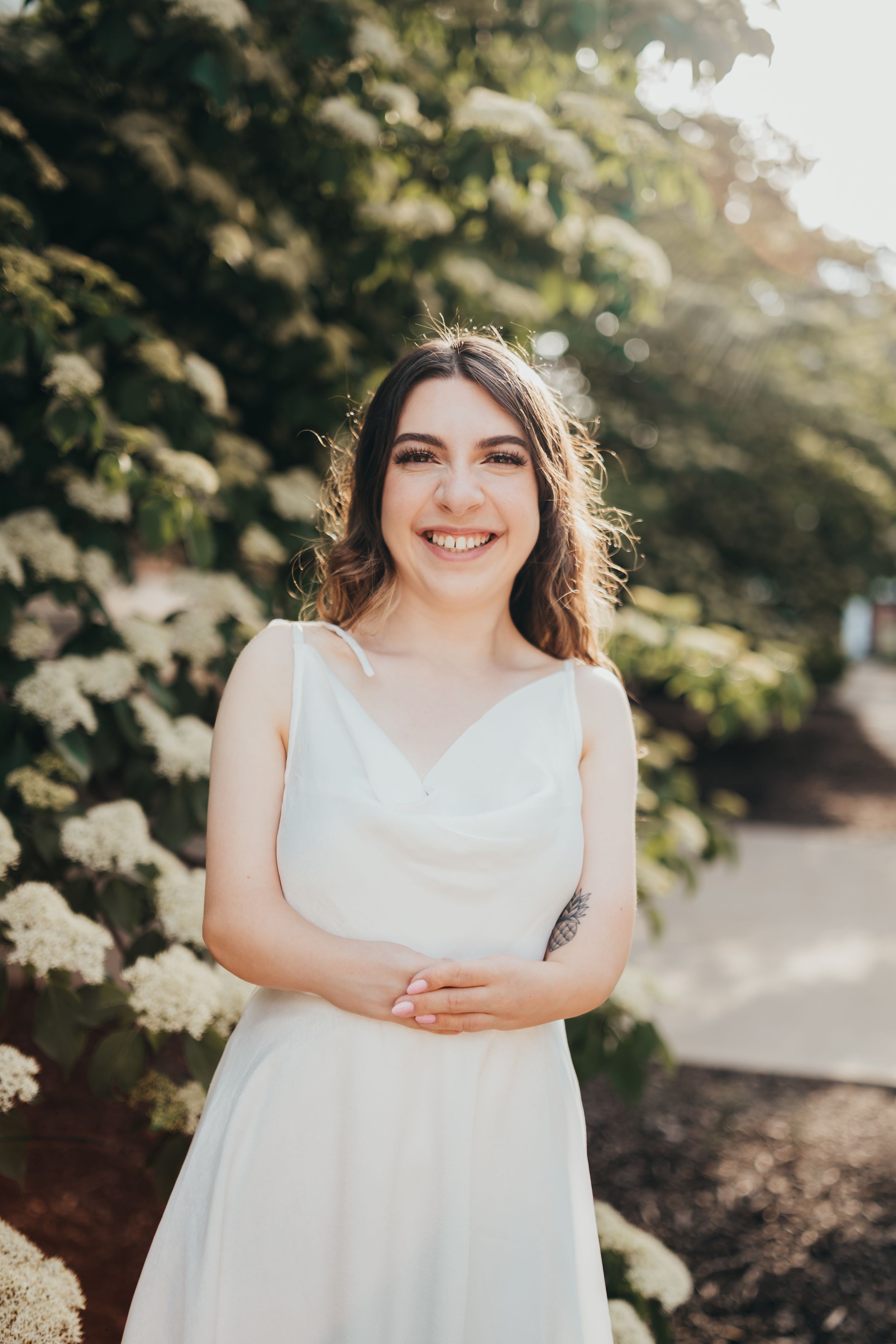 Person in white dress posing outdoors near flowering bushes.