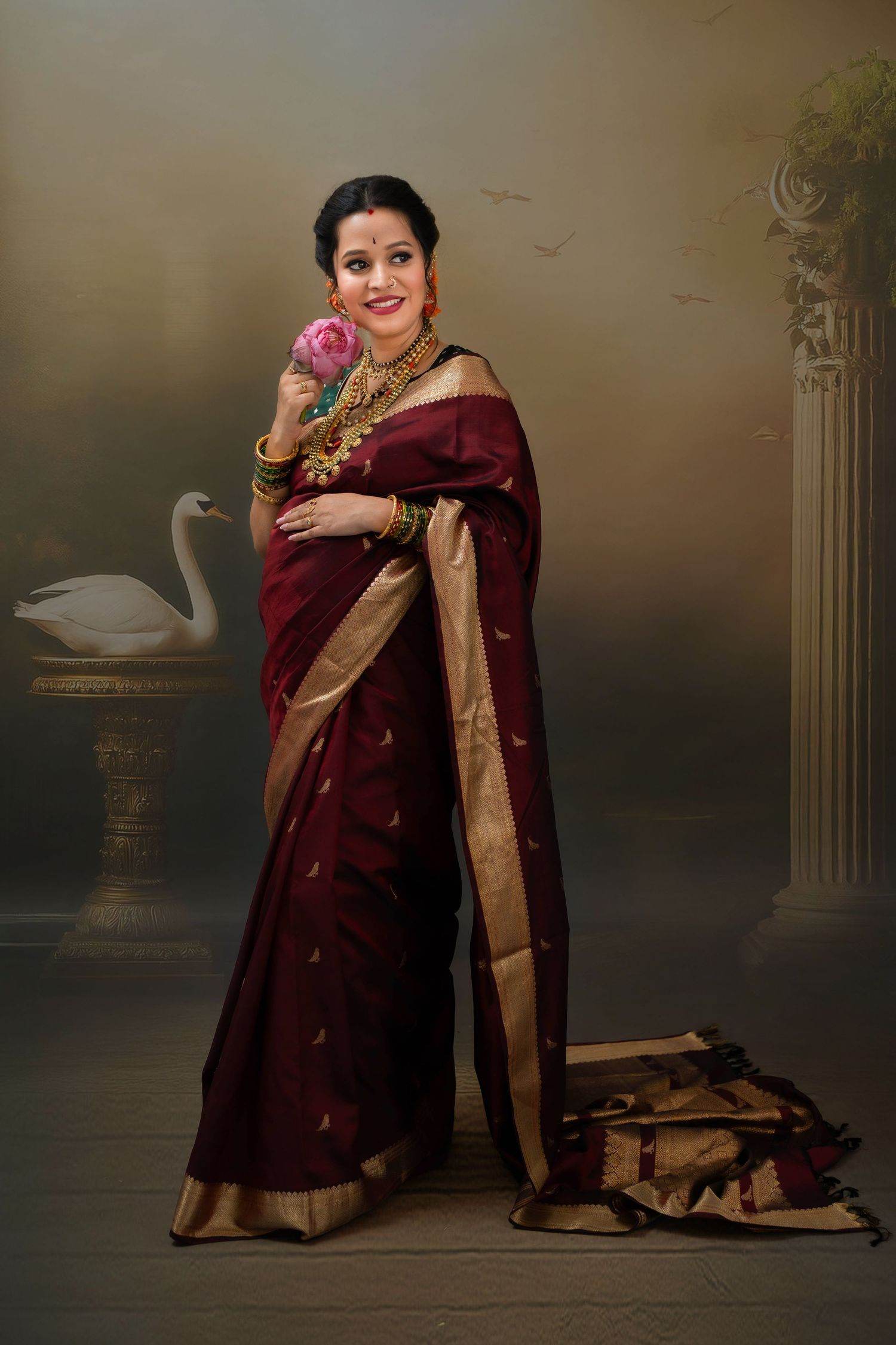 Pregnant woman in maroon silk saree holding her baby bump during a calm studio maternity photoshoot, capturing a quiet moment of motherhood.