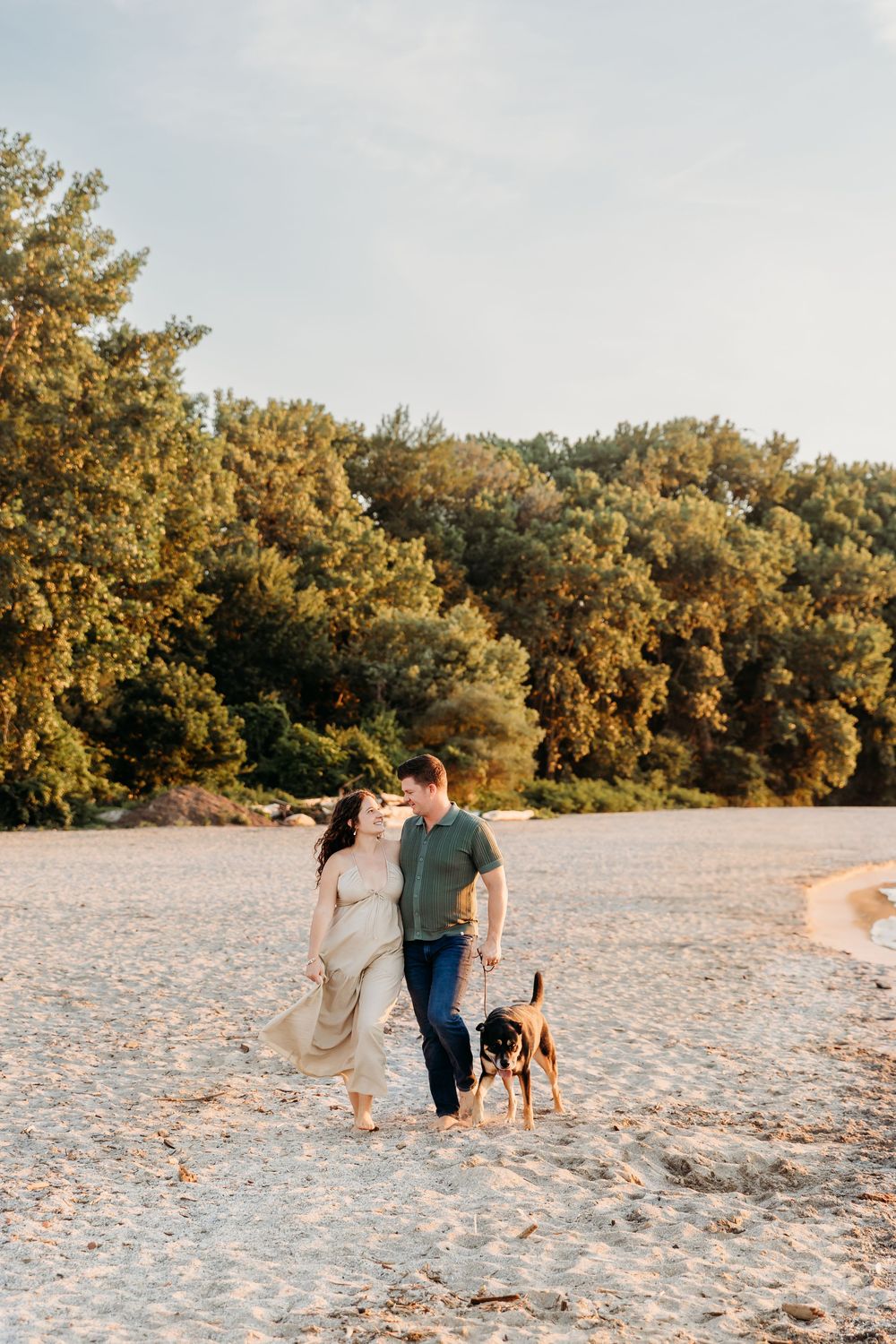 A couple walks with their dog on a sandy beach at sunset with trees in the background.