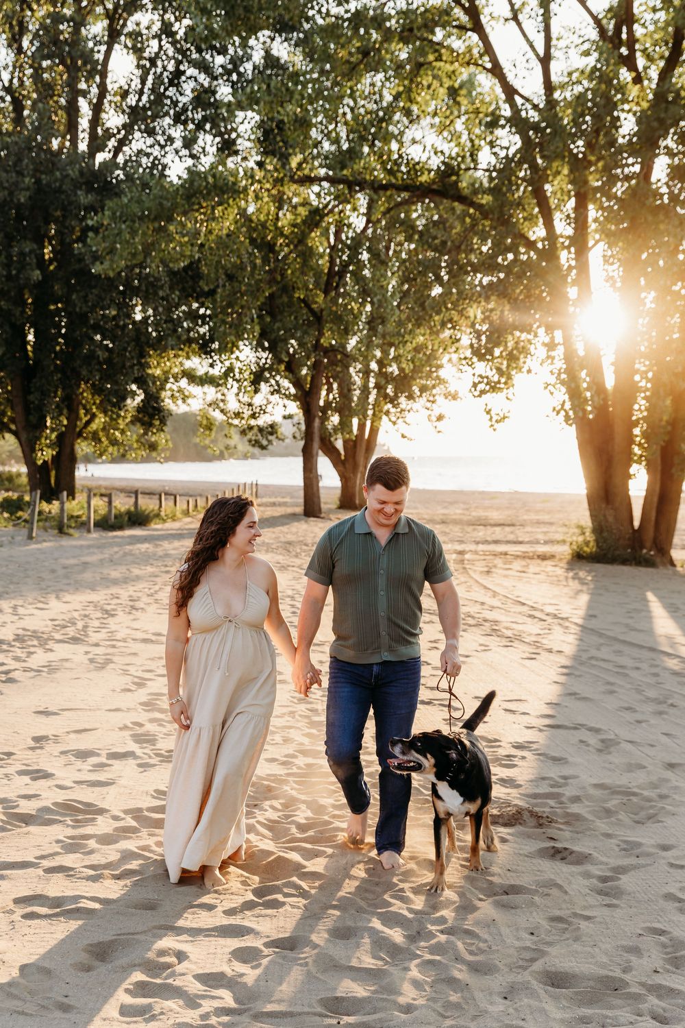 A couple walks together on a sandy beach at sunset while golden sunlight streams through trees. Cleveland Maternity Photographer
