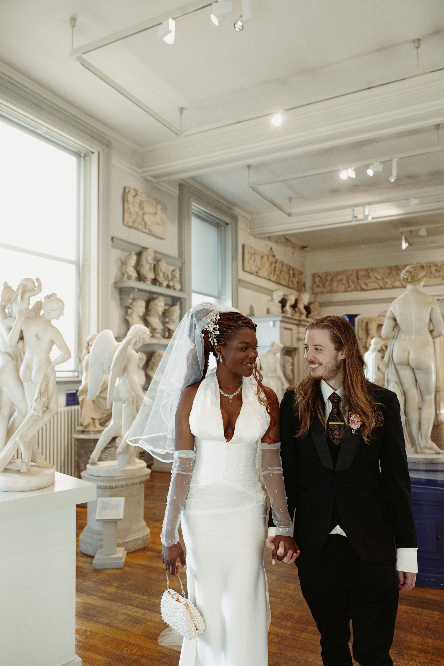 Two people walk through an art gallery displaying classical marble sculptures and statues against white walls.