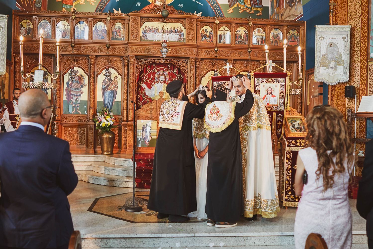 Orthodox wedding ceremony with priests holding crowns over couple at ornate golden iconostasis altar.