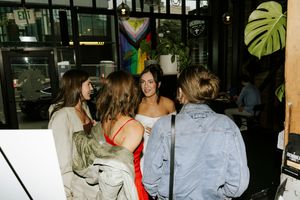 A group of people gather in a dimly lit indoor space with monstera plants visible in the background.
