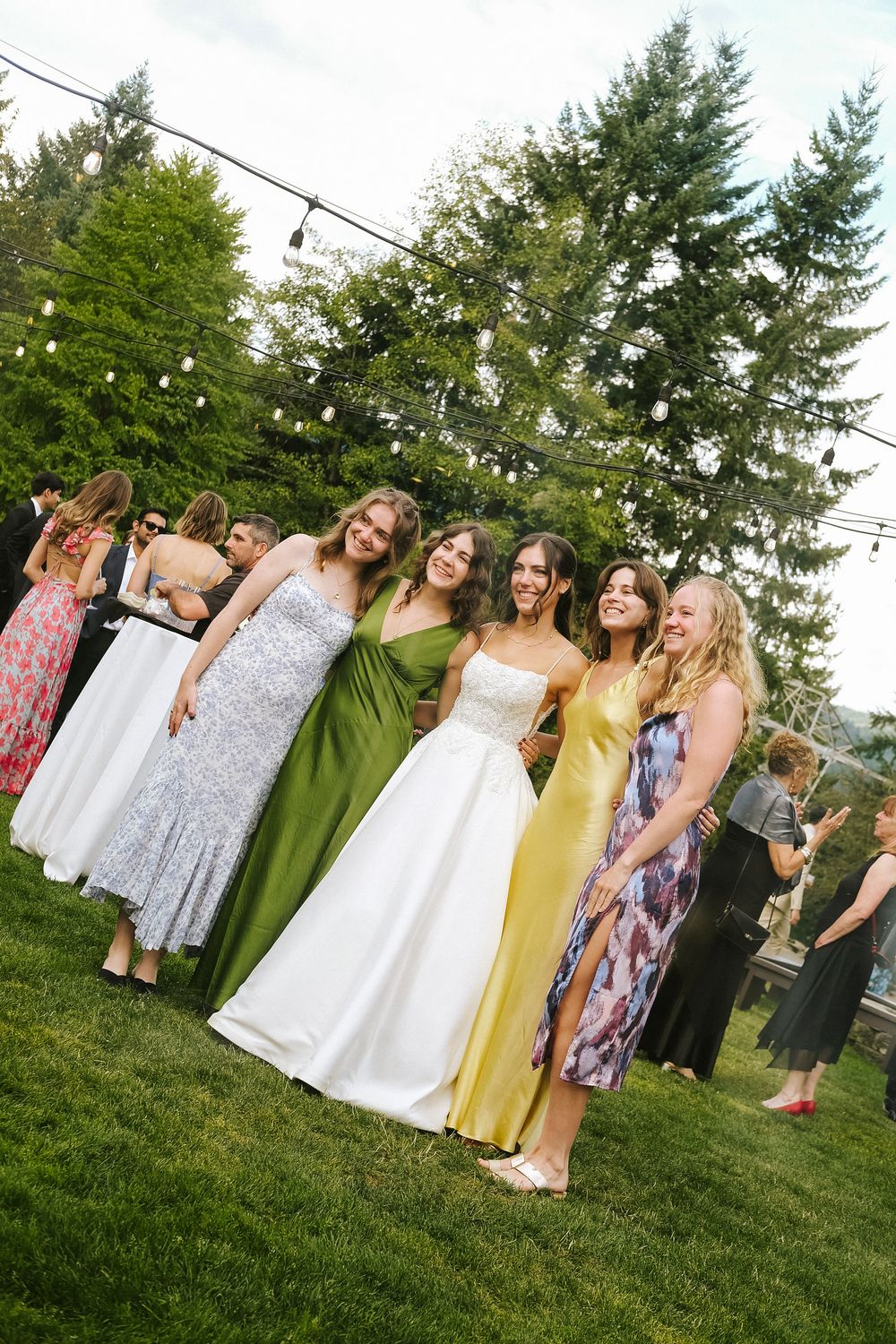 A group of wedding guests in colorful dresses gather on a green lawn during an outdoor summer celebration.