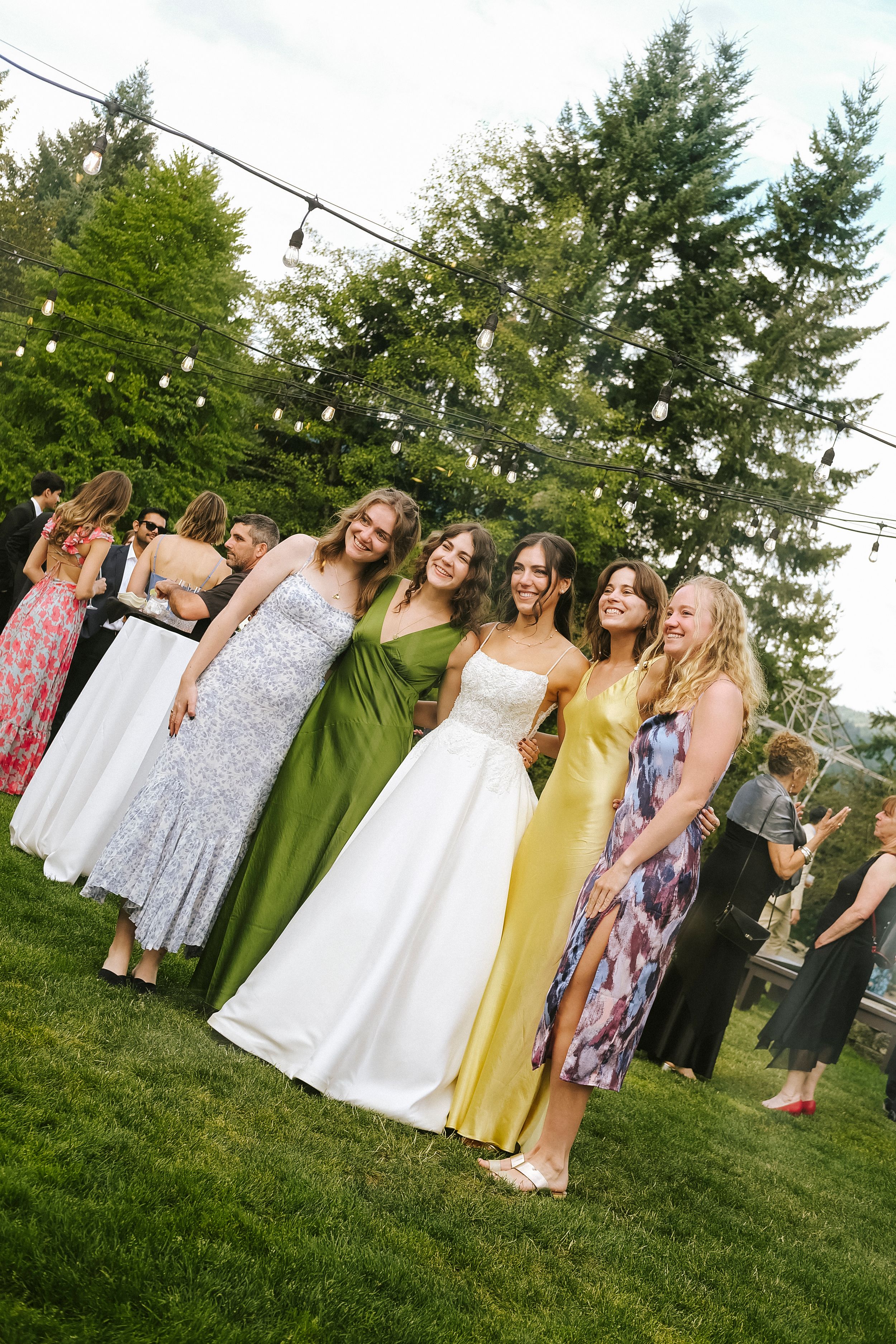 A group of wedding guests in colorful dresses gather on a green lawn during an outdoor summer celebration.