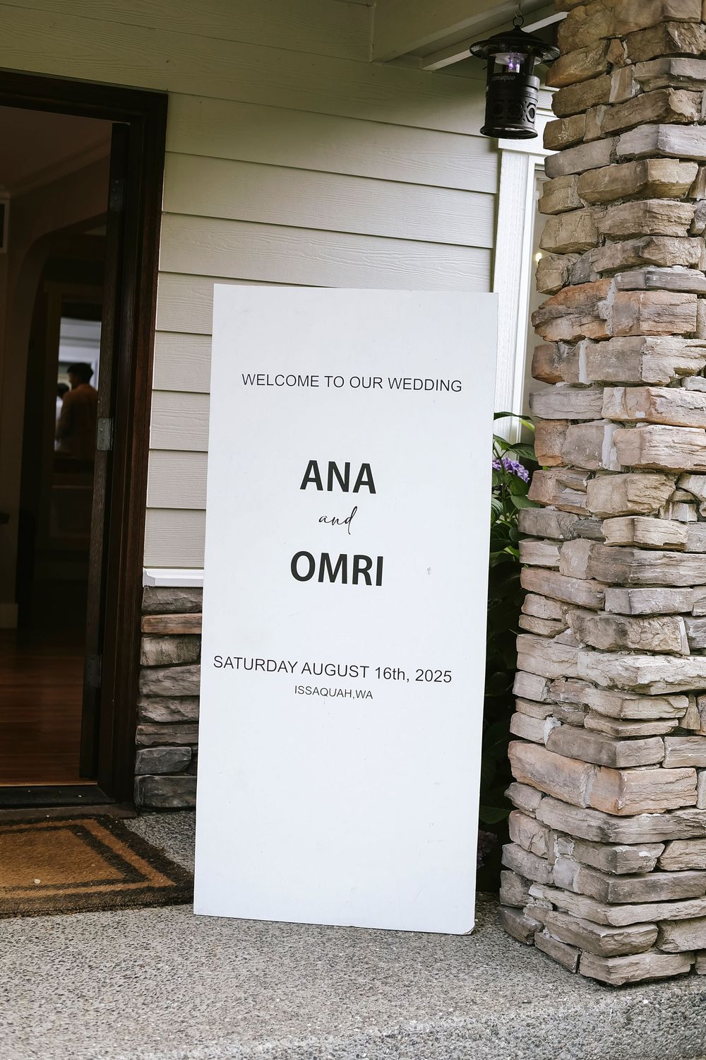 A minimalist white wedding welcome sign for Ana and Omri's ceremony displayed against a stone pillar entrance.