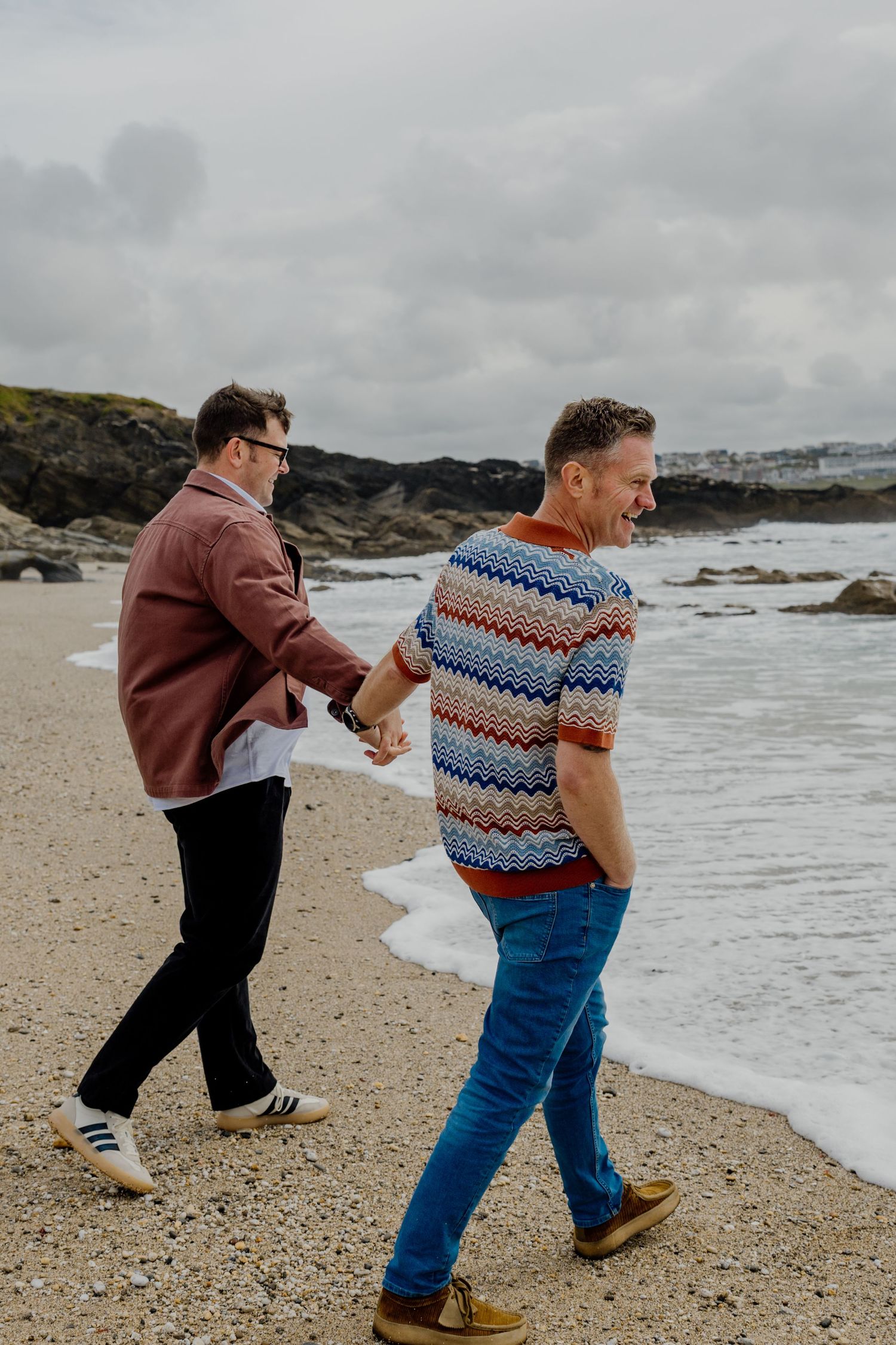 Two men holding hands while walking on a sandy beach with ocean waves and a coastal town in the background.