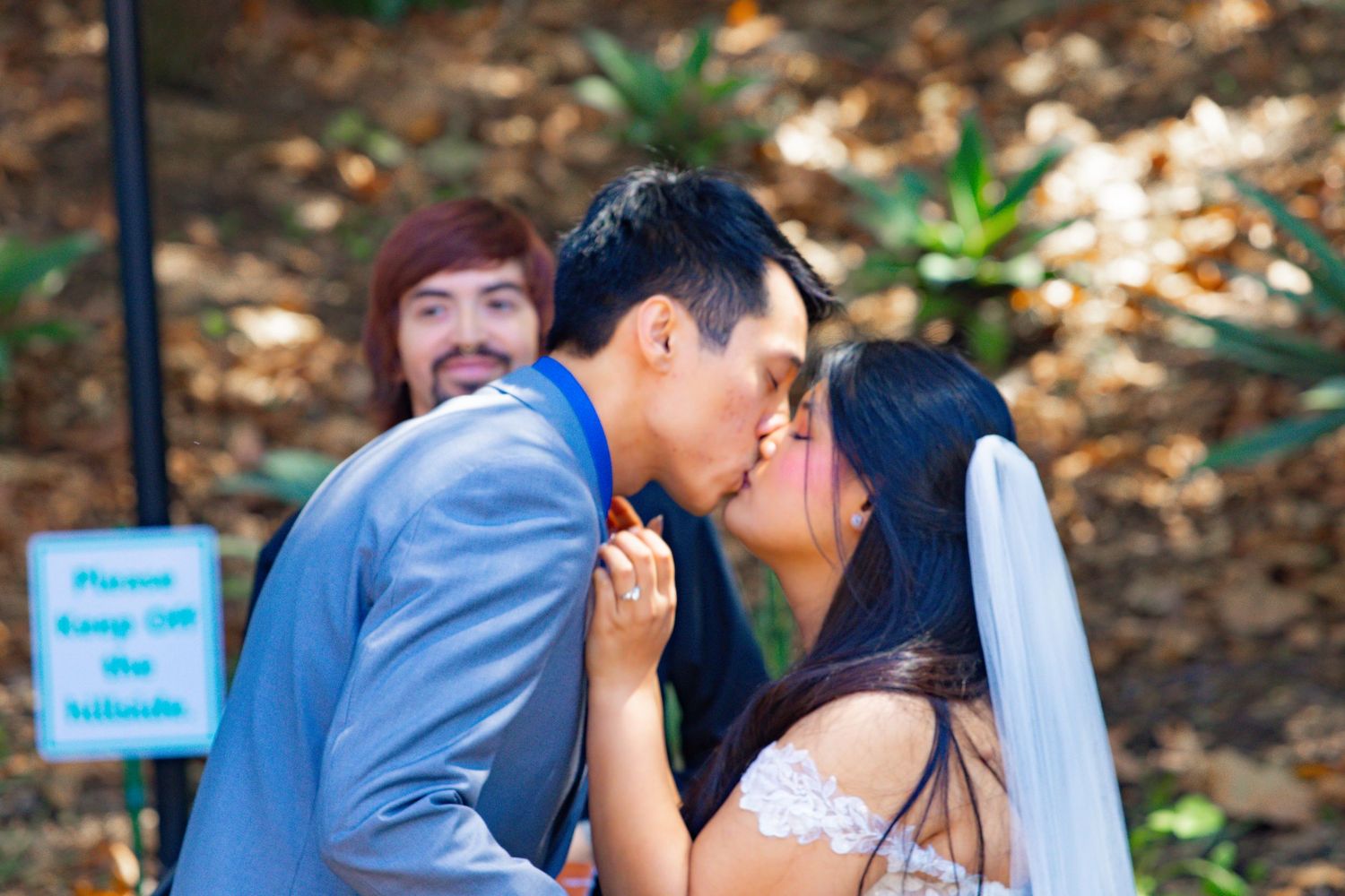 Bride and groom share a first kiss at outdoor wedding ceremony while guest photobombs smiling in background.