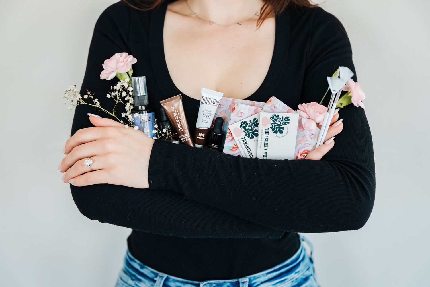 Woman in black top holding beauty products and flowers crossed over her chest against a gray background. Facials and holistic skincare.