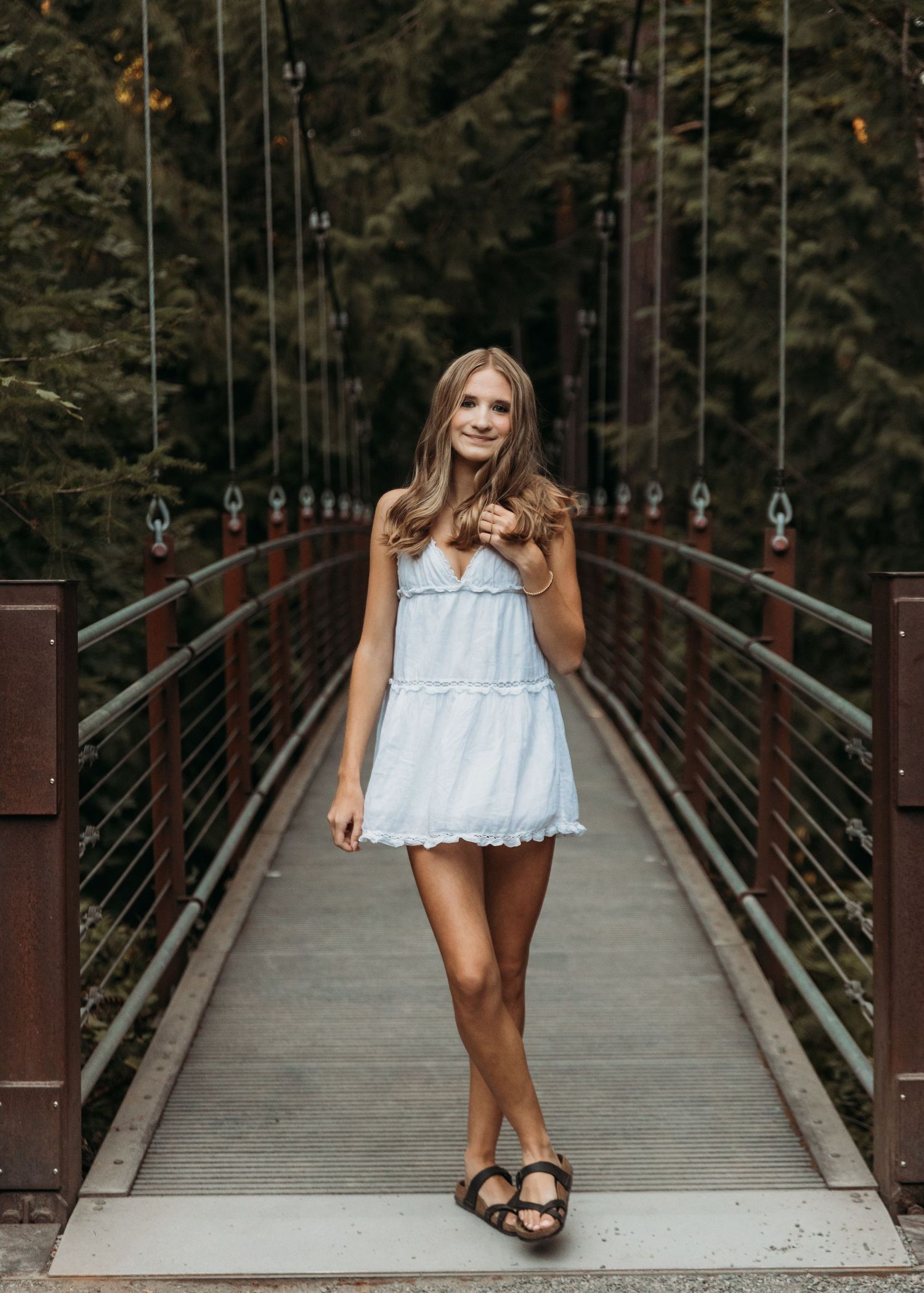 Young woman in light blue mini dress and sandals smiling on a wooden suspension bridge surrounded by green trees.