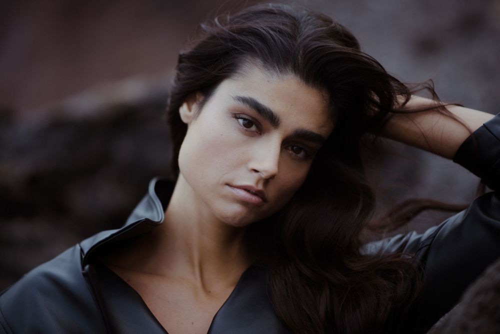 Young woman with dark hair posing outdoors in a black leather jacket, looking intensely at the camera.