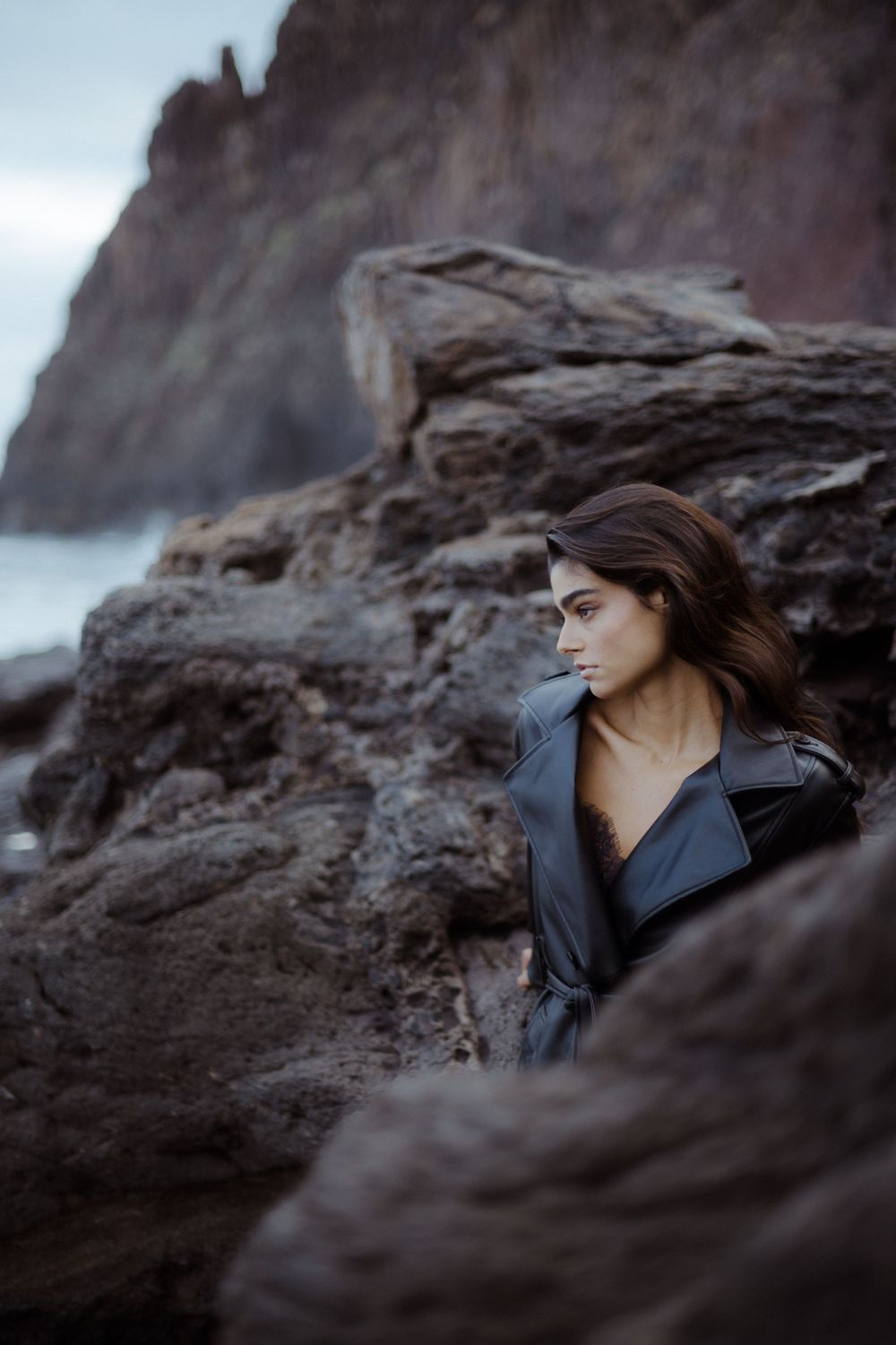 A young woman in a black leather jacket poses among rugged coastal rocks with dramatic cliffs in the background.