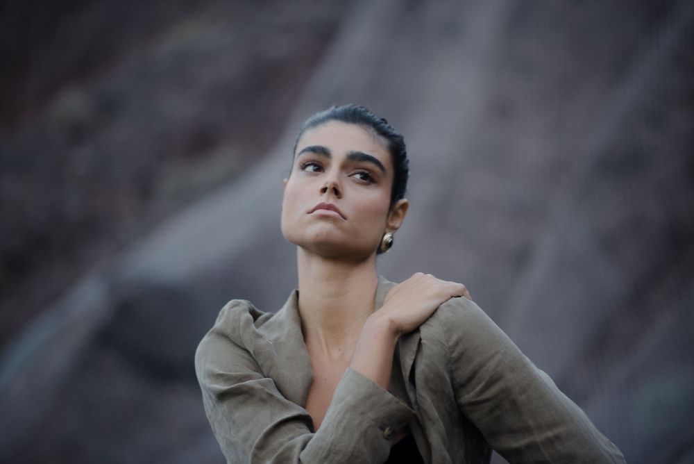 A young woman in an olive jacket gazes upward, posing against a rocky mountain backdrop.