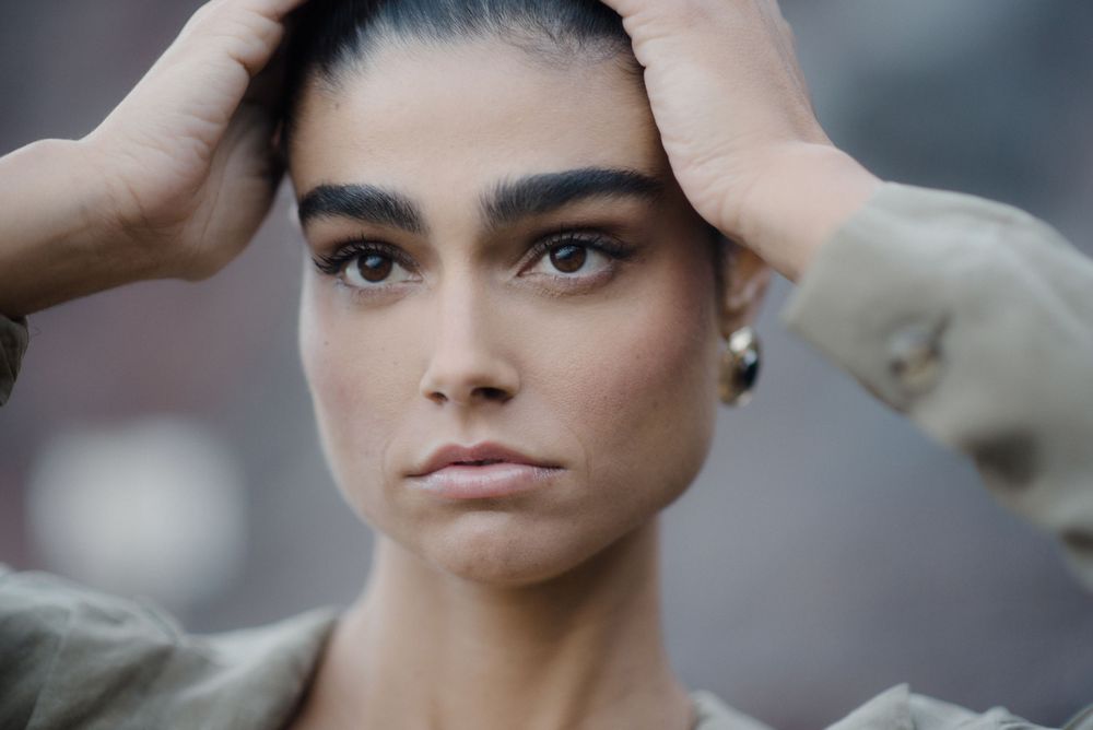 Close-up portrait of a young woman with bold brows, holding her hair back, wearing a green jacket, serious expression.
