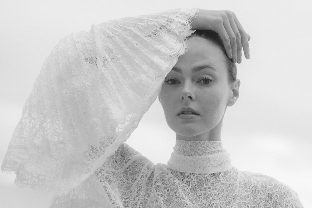 Black and white portrait of a woman in white lace high-neck dress holding a lace veil overhead.
