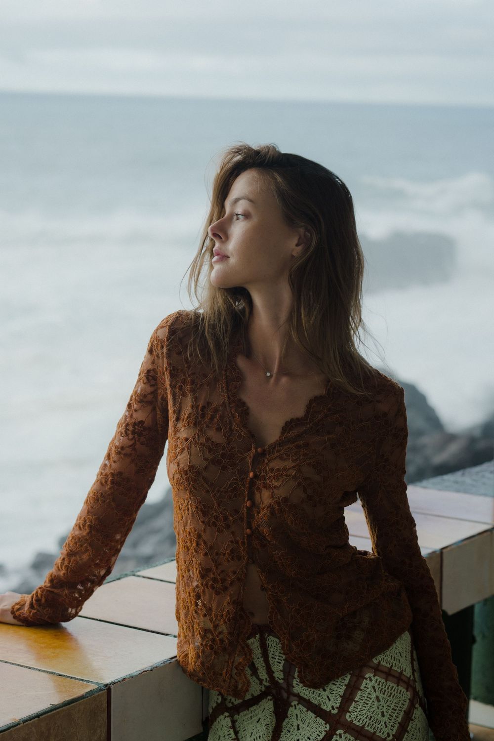 Woman in brown lace top gazes at ocean waves from a seaside balcony, hair blowing in the breeze.