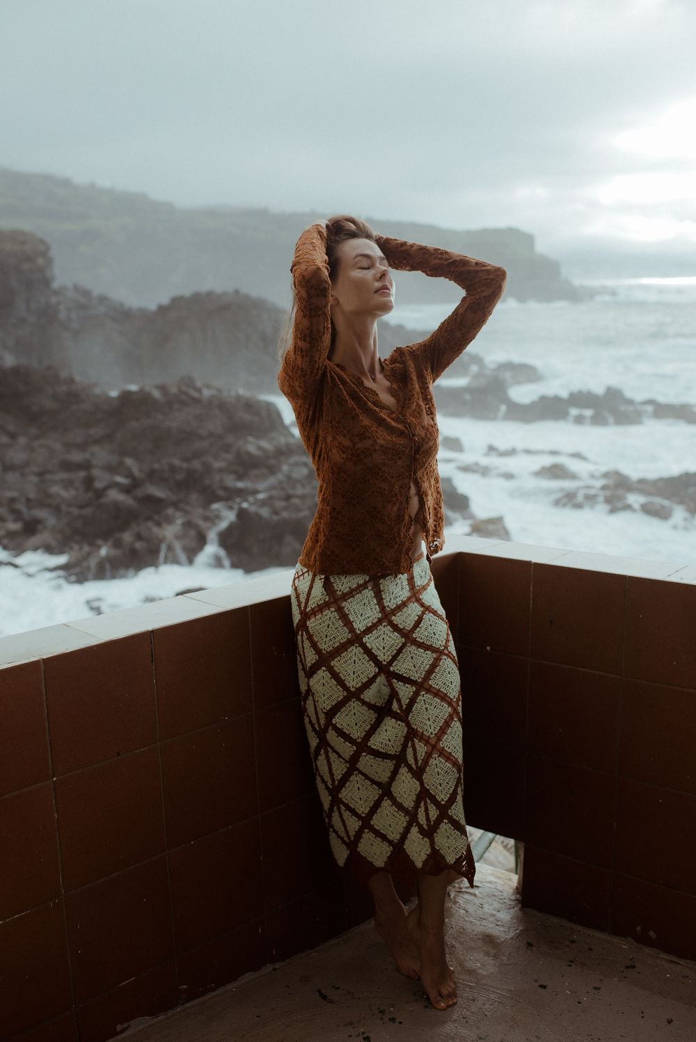 Woman in brown knit sweater and crochet skirt poses on coastal balcony with dramatic ocean waves behind her.