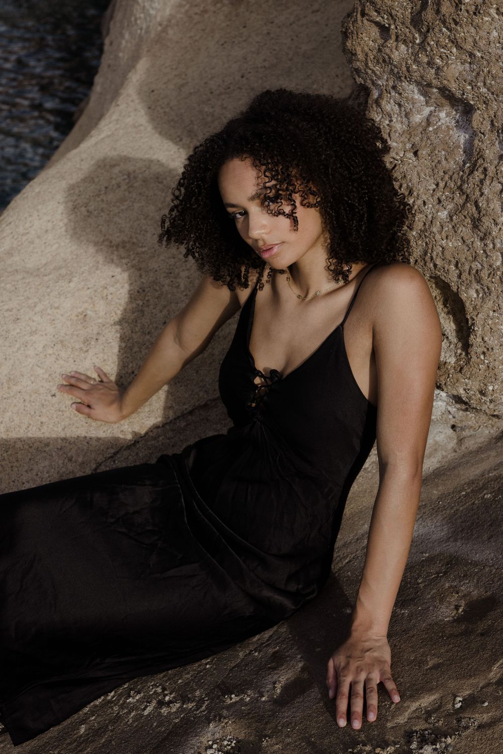 A woman with curly hair in a black slip dress poses gracefully against large rocks outdoors.