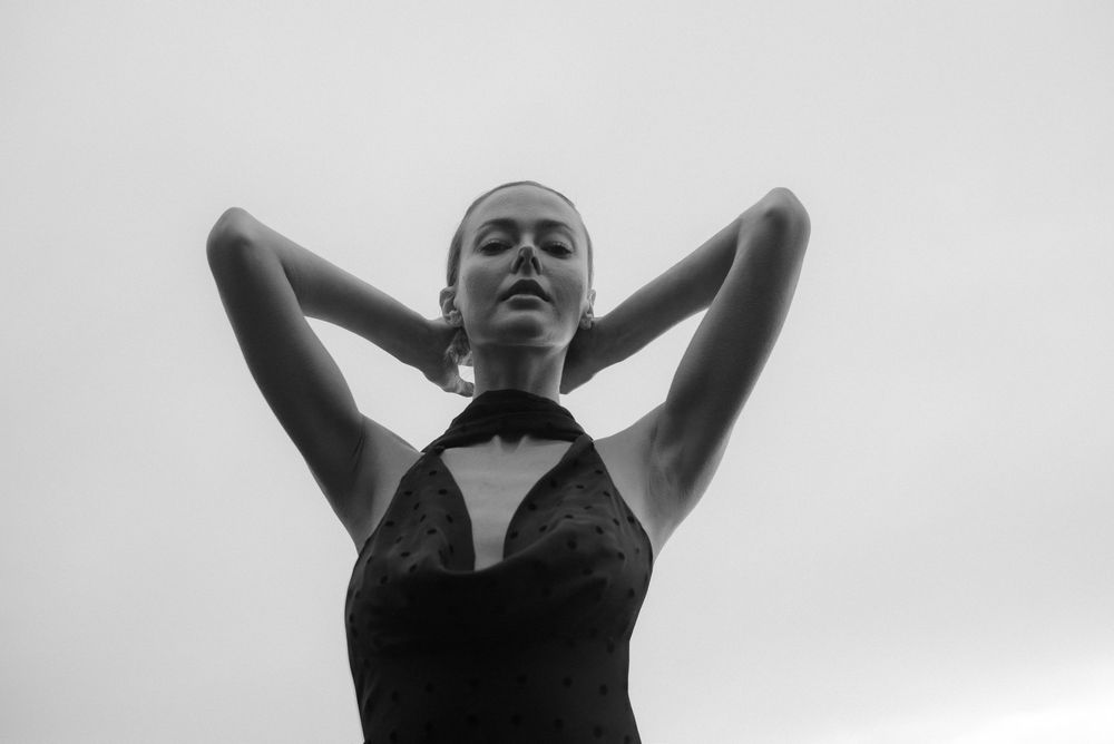 Black and white photo of a woman in a black dress with arms raised behind her head against a grey sky.