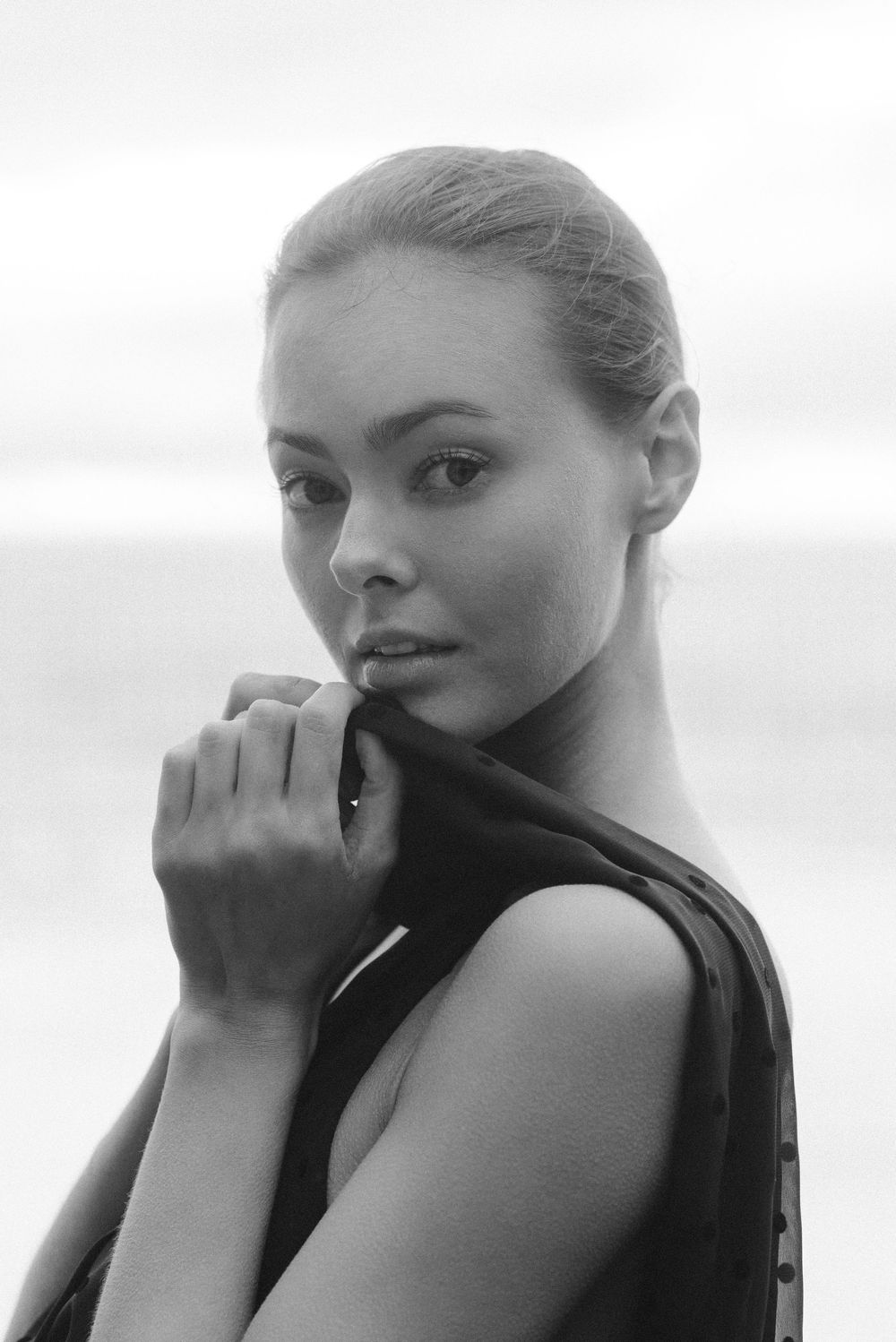 Black and white portrait of a young woman holding dark fabric, gazing at camera with ocean background.