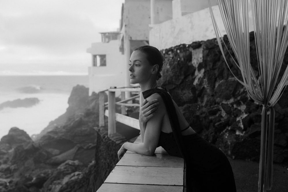 Woman in black dress gazes at rocky ocean coastline from a balcony in a moody black and white photo.