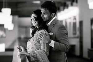 Black and white photo of a couple embracing, woman in embroidered dress, man in suit, smiling together indoors.