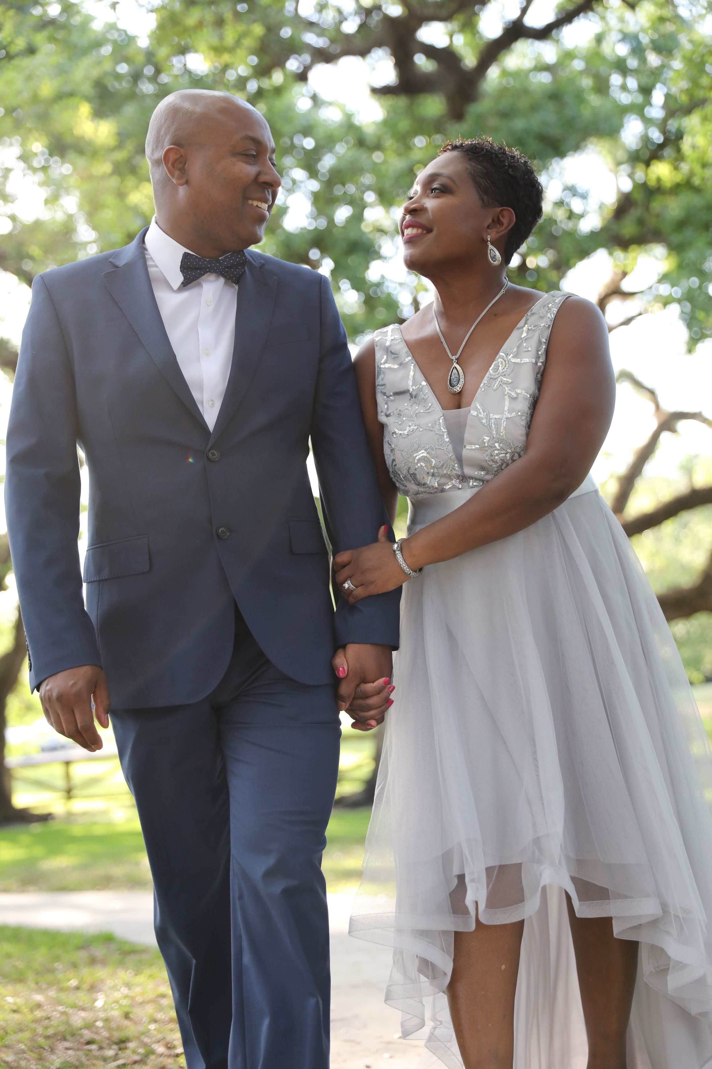 A bride in a flowing white dress and groom in navy suit share a loving moment under trees.