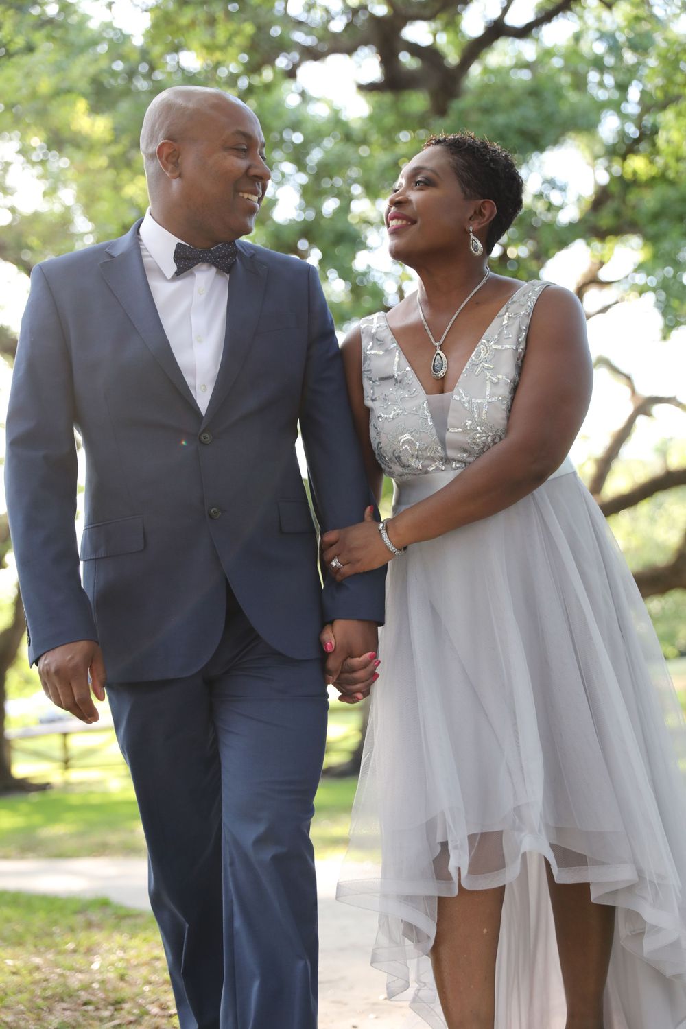 A bride in a flowing white dress and groom in navy suit share a loving moment under trees.