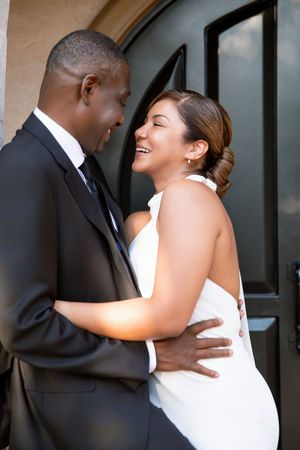 A joyful couple in wedding attire embraces and laughs together in front of elegant black doors.