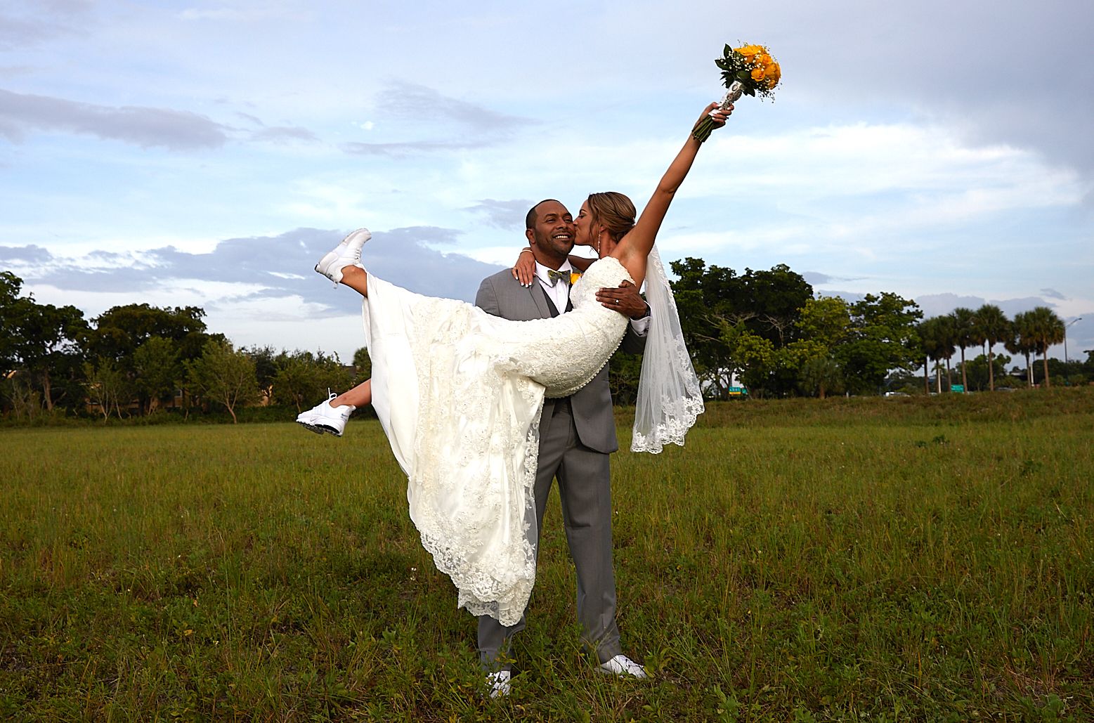 A playful wedding photo of a groom lifting his bride in a white dress against a sunset sky.