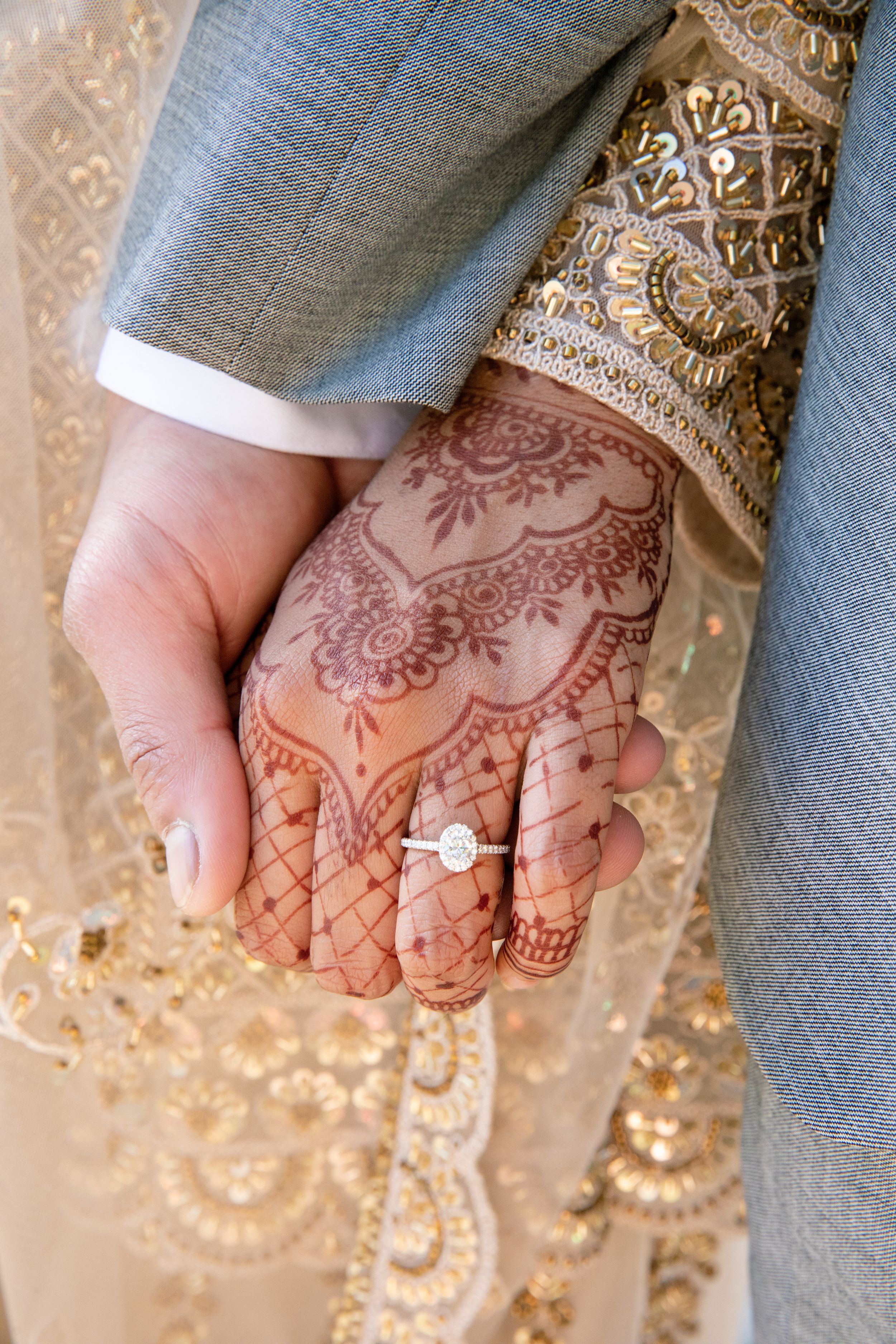 Bride and groom holding hands, bride adorned with intricate henna mehndi design and diamond ring, wearing gold embroidered attire.