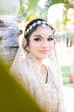 South Asian bride in white and gold embroidered lehenga with maang tikka and veil poses outdoors.
