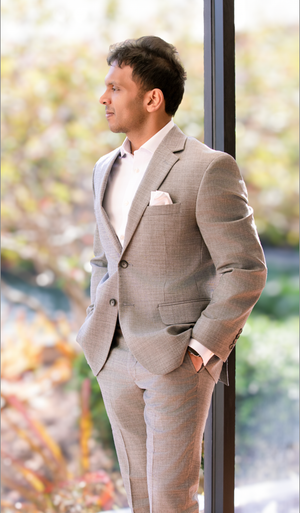 A man in a tailored light gray suit with pocket square stands by a window, gazing outdoors at autumn foliage.