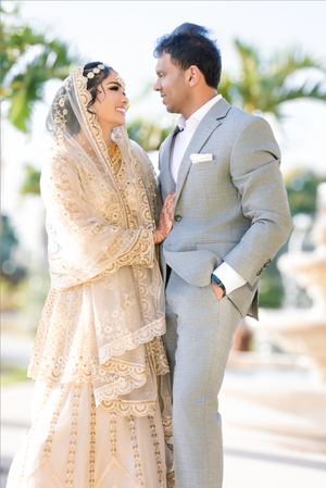 South Asian bride in ivory lace dupatta and groom in gray suit share a romantic moment outdoors on their wedding day.