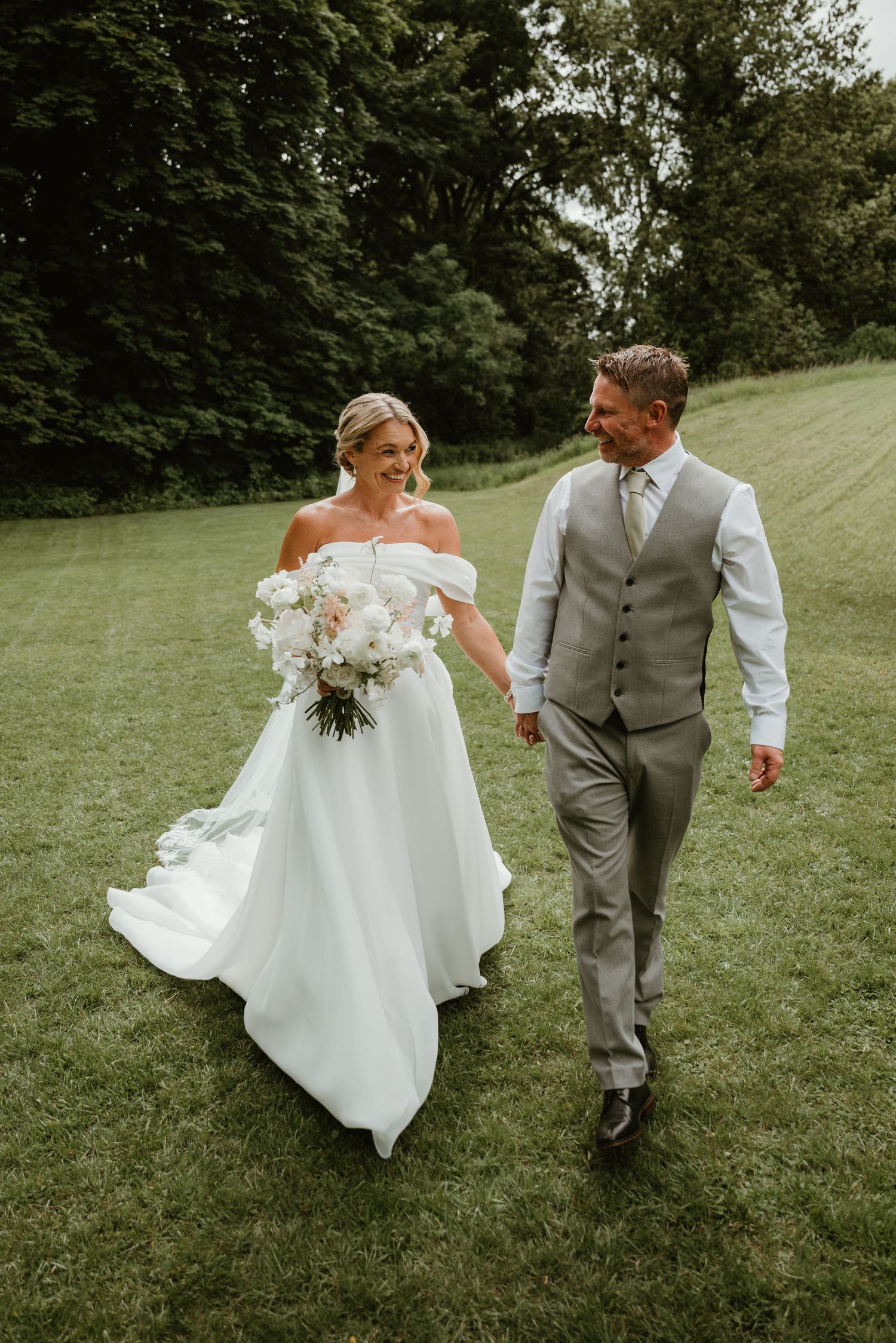 Bride in off-shoulder gown holding white bouquet walks hand-in-hand with groom in gray suit on lush green lawn.