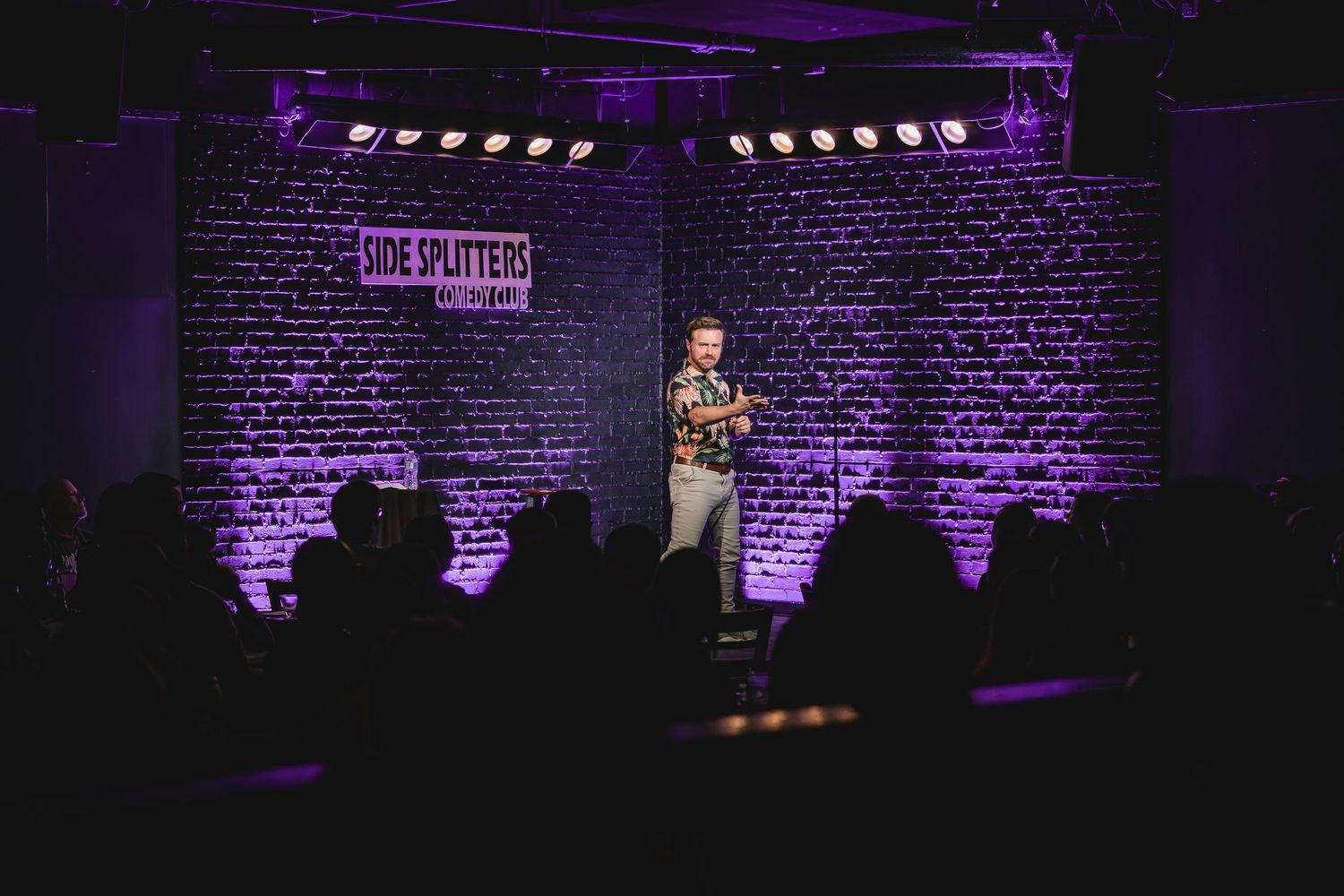 A stand-up comedian performs on stage at a comedy club with a brick wall and partial Comedy Bone sign behind him.