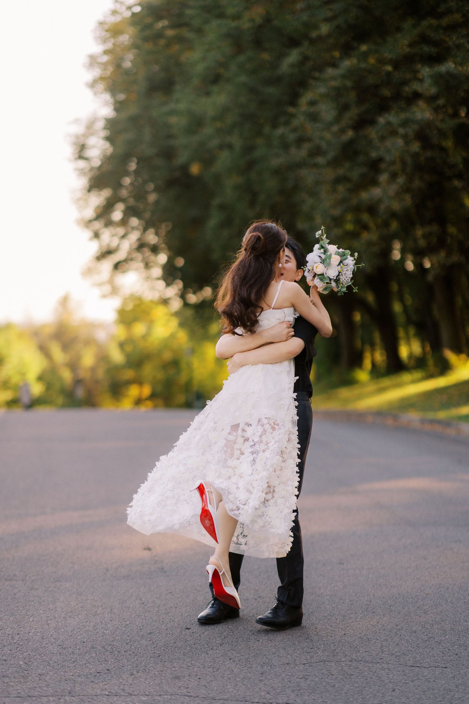Groom carries bride in white dress as they kiss on a tree-lined road during golden hour wedding photo.