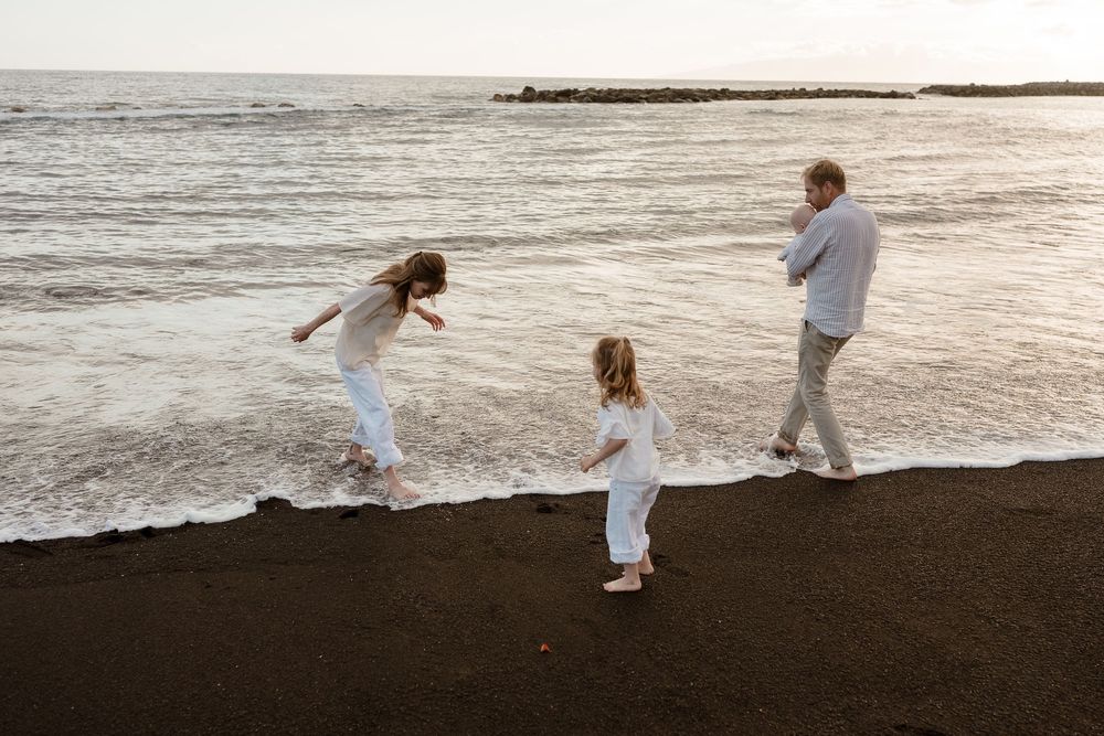 Family of three playing on a black sand beach at sunset, wearing white outfits near ocean waves.
