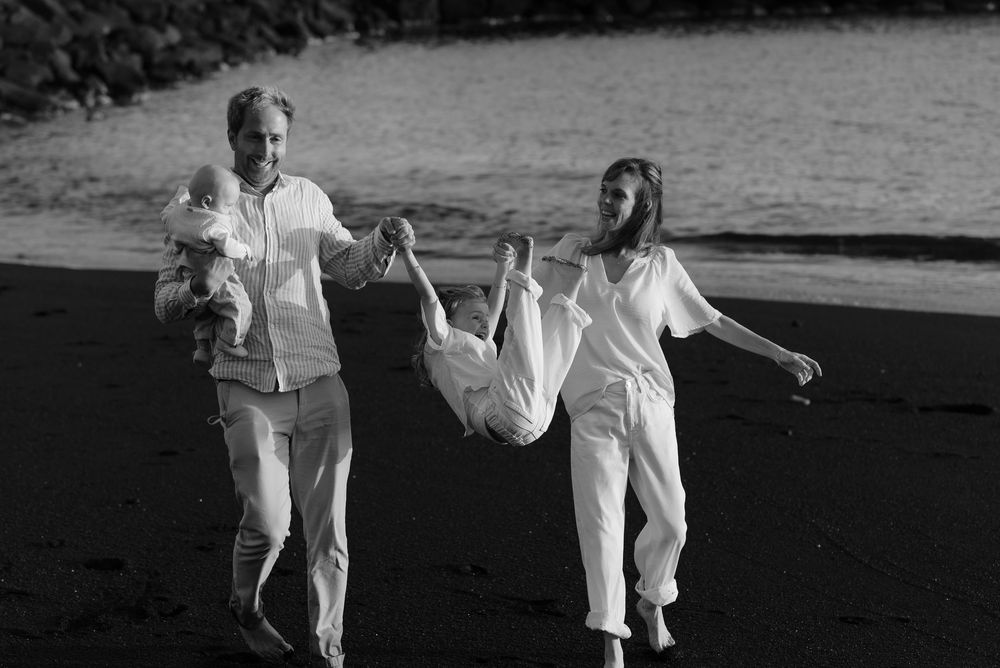 Black and white photo of a family on a dark sandy beach, parents swinging child between them.