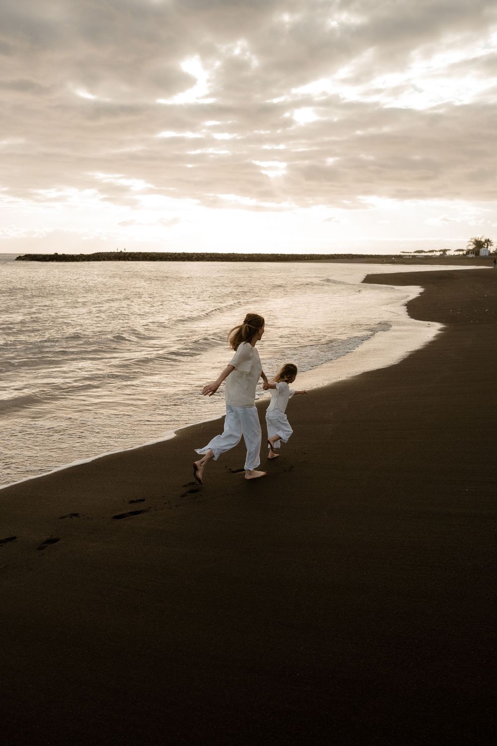 Mother and child in white running along black sand beach at golden sunset.