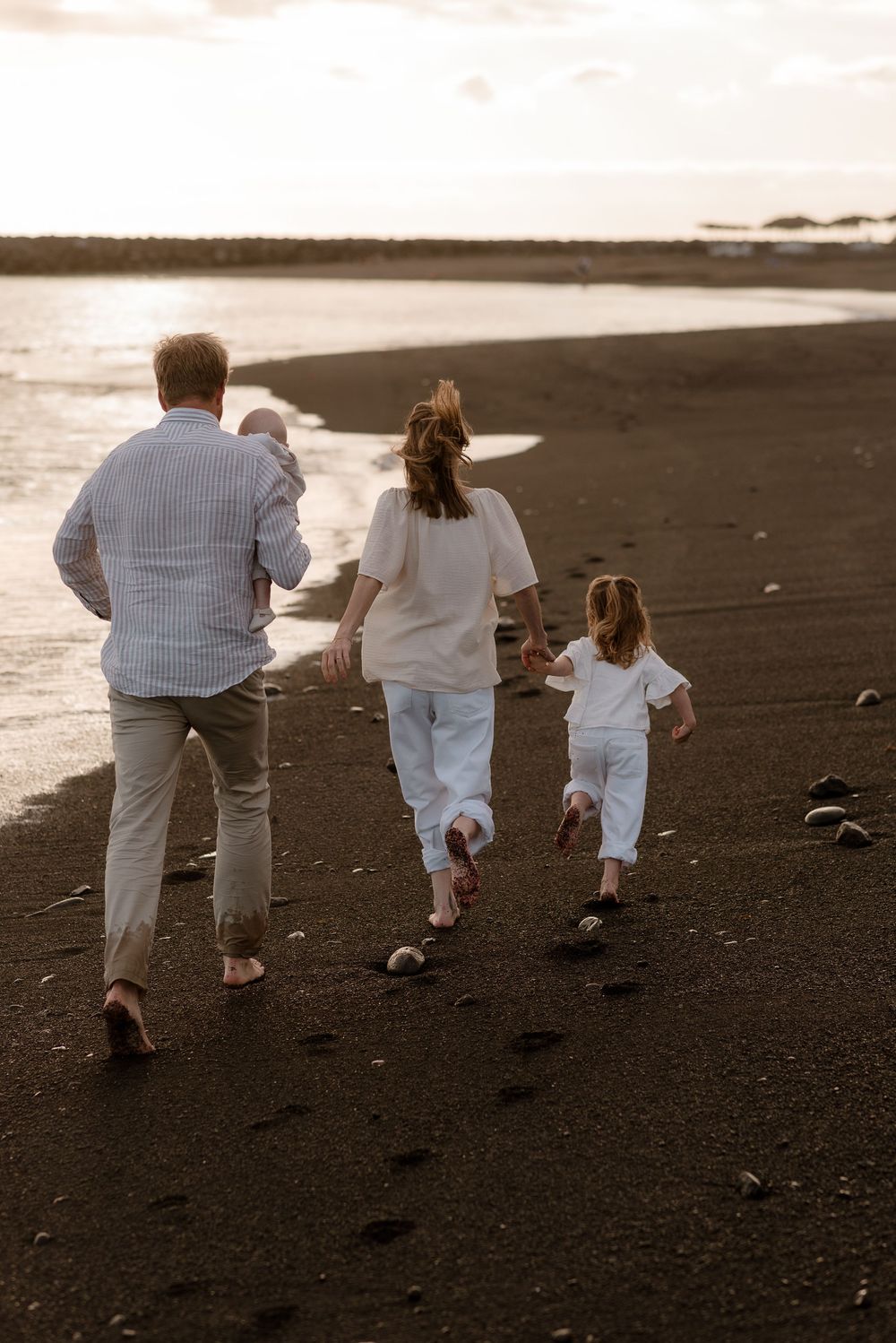 Family walking barefoot on black sand beach at sunset, parents with two young children enjoying a peaceful seaside stroll.