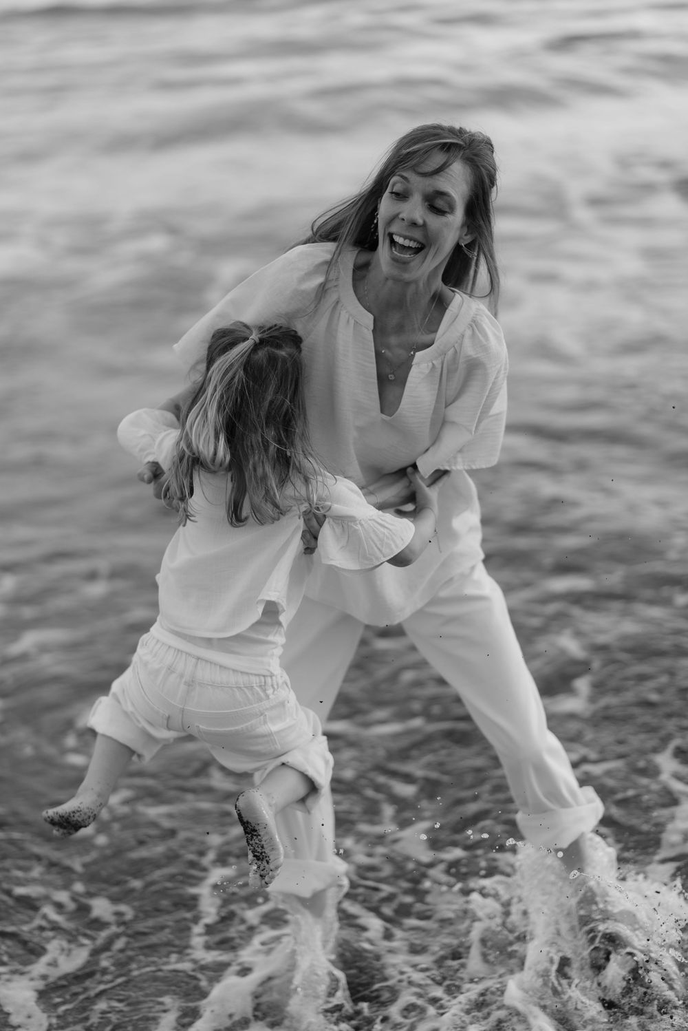 Black and white photo of laughing mother hugging young daughter in ocean waves.