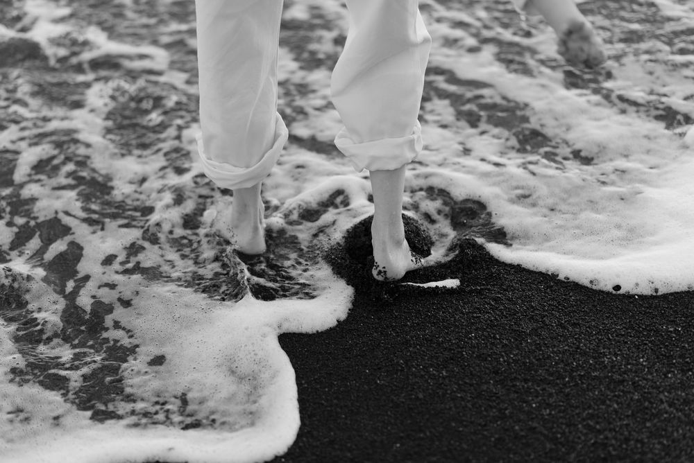 Black and white photo of person standing barefoot in ocean waves on dark sand beach.