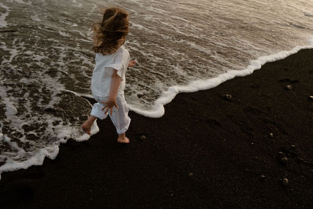 A young girl in a white dress walks along a black sand beach as ocean waves wash over her bare feet.