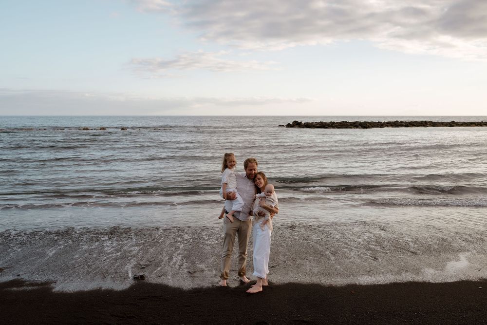 Family of three posing on a black sand beach with ocean waves and cloudy sky backdrop.