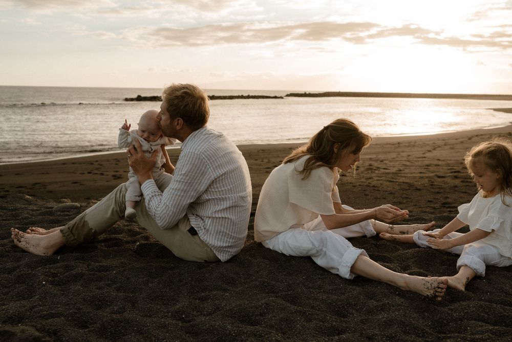Family sitting together on a sandy beach at sunset, parents with young children enjoying a peaceful seaside moment.