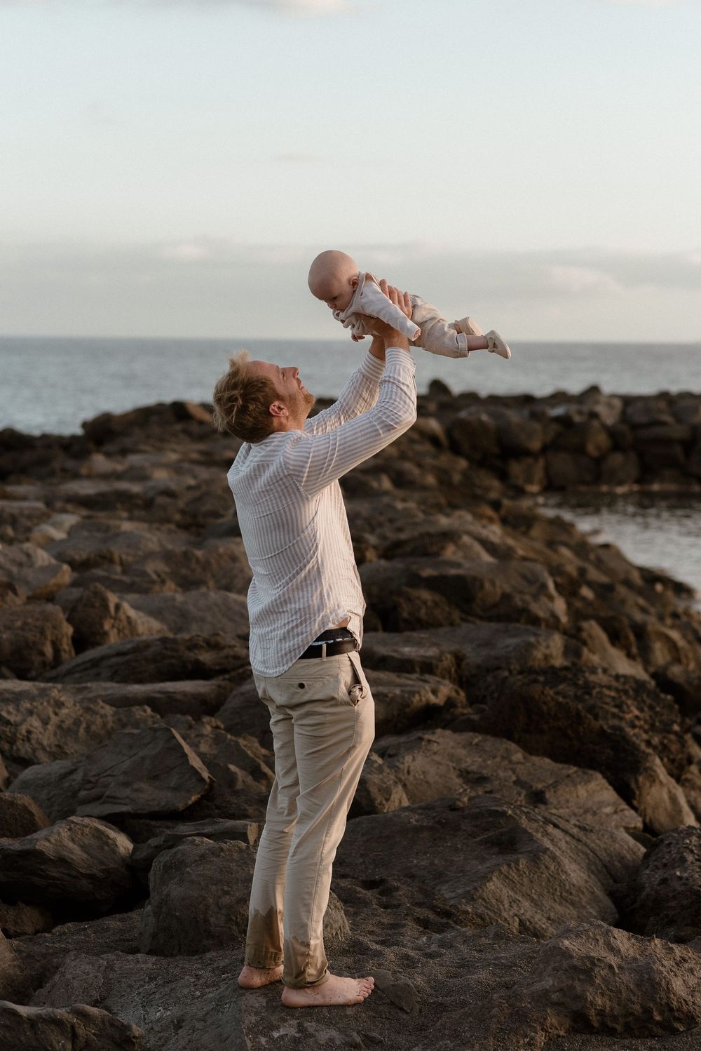 Father lifting baby joyfully overhead while standing on rocky coastal shore at dusk near the ocean.